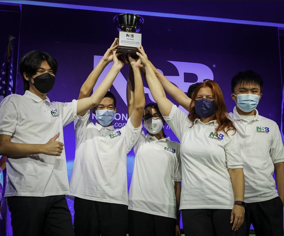 The team in National Science Bowl polos holding a trophy on stage, wearing masks